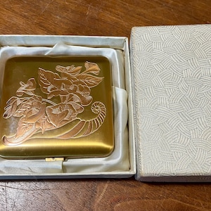 May include: A gold-toned powder compact with an embossed floral design in a matching box. The compact is square with a hinged closure. The box is off-white with a textured pattern and the words "POWDER CASE" printed on it.