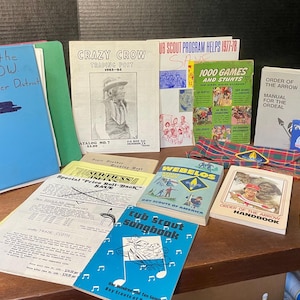 May include: A collection of vintage Boy Scouts of America literature and ephemera, including a blue binder labeled Order of the Arrow, a Crazy Crow trading post catalog, a 1000 Games and Stunts book, a Webelos manual, a songbook, and a Scout Handbook.