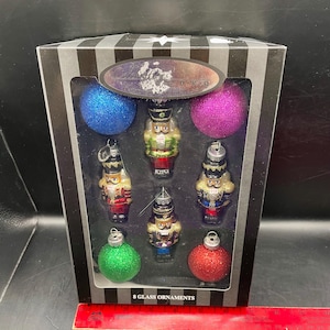 May include: A boxed set of eight glass Christmas ornaments. The set includes six miniature nutcracker figurines and two glittery ball ornaments in blue, green, purple, and red. The box has a black and white striped design and the text "8 GLASS ORNAMENTS".