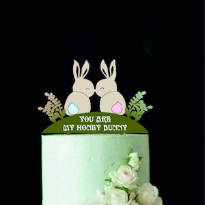 Op de afbeelding: Een taarttopper met twee konijnen die elkaar kussen met de tekst "You are my honey bunny" op een groene basis met bloemmotieven.