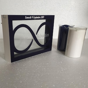 May include: A white wooden frame with a navy blue border and a glass infinity symbol inside. The frame has the text "Samedi 4 Septembre 2021" and "Sébastien" inscribed on it. Two glass containers, one blue and one white, are next to the frame.