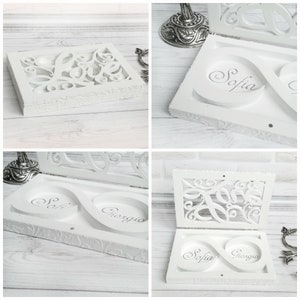 May include: White wooden ring box with an infinity symbol design. The box is open and the two halves are separated. The top half has a cutout design of the word "love". The bottom half has two compartments with the names "Sofia" and "Giorgio" inscribed inside.