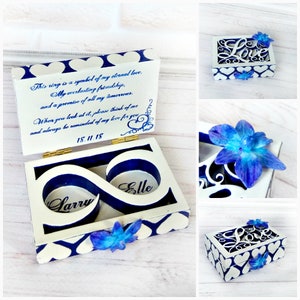 May include: White wooden ring box with blue infinity symbol design. The box has two compartments for rings, labeled "Larry" and "Elle". The lid has a blue and white heart pattern and a message that reads: "This ring is a symbol of my eternal love, my everlasting friendship, and a promise of all my tomorrows. When you look at it, please think of me and always be reminded of my love for you. 18.11.18"