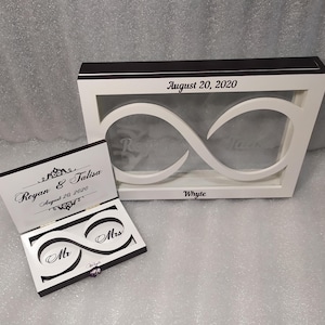 May include: White wooden box with a clear infinity symbol cutout. The box has the text "August 20, 2020" and "Whyte" inscribed on it. A smaller white wooden box with a black trim and an infinity symbol cutout is open and has the words "Mr" and "Mrs" inscribed on it. The smaller box has the text "Royan & Talisa August 20, 2020" inscribed on it.