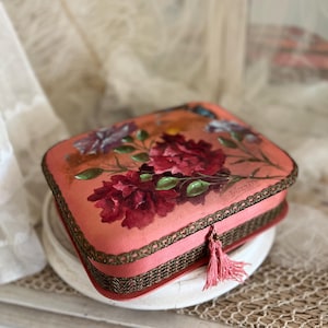 May include: A rectangular, coral-colored box with a floral design on the lid. The box features a dark red trim, a decorative gold border, and a pink tassel. The floral design includes burgundy and purple flowers with green leaves.