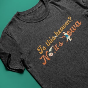 以下が含まれることがあります： 暗いグレーのTシャツに、ヴィンテージスタイルのグラフィックがプリントされています。グラフィックには「Is this heaven? No, it's Iowa」という文字と、野球選手のイラストが描かれています。