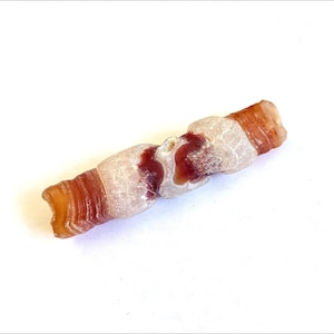 May include: A cylindrical stone bead featuring a banded pattern of translucent white and deep reddish-brown layers. The surface displays a natural, uneven texture with distinct color transitions and a polished, semi-glossy appearance throughout.
