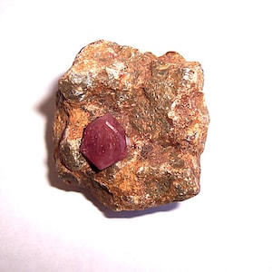 May include: A red ruby crystal embedded in a rough, brown rock. The ruby is a hexagonal shape with a smooth, shiny surface.