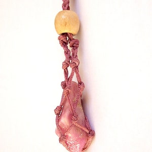 May include: A pink crystal is wrapped in a pink macrame cord and hangs from a wooden bead. The crystal is a light pink color and has a smooth, polished surface.