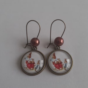 May include: A pair of bronze earrings with brown beads and round glass charms featuring a vintage illustration of a family of rabbits.