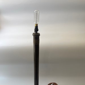 May include: A vintage table lamp with a dark brown metal stem and base. The lamp has a decorative label and a clear, elongated light bulb. The base is a dark wooden block. A brown, braided power cord is attached. The lamp's height is approximately 40 cm.