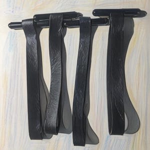 May include: Four black leather straps with metal clasps hanging against a light background with a textured pattern.