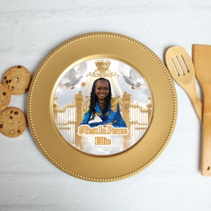 Puede incluir: Un plato de servicio dorado con un centro blanco que presenta una foto de una joven con un atuendo azul y dorado, una corona y un par de palomas blancas. El texto "Rest In Peace Ellie" está impreso debajo de la foto.