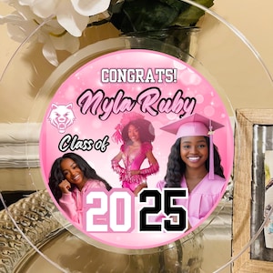 以下が含まれることがあります： 狼のマスコット、テキスト「Congrats! Nyla Ruby Class of 2025」、卒業生の3枚の写真が描かれたピンクと白の卒業パーティー用プレートです。