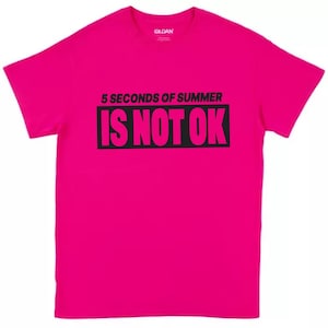 May include: A bright pink t-shirt with the text "5 SECONDS OF SUMMER IS NOT OK" in black. The shirt is a crew neck style and appears to be made of cotton. The Gildan brand logo is visible on the upper back of the shirt.