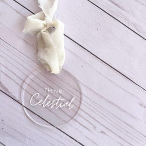 May include: A clear round acrylic ornament with the text "Think Celestial" written in white cursive script. The ornament is hanging from a white ribbon.