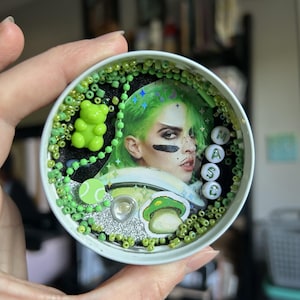 Pode incluir: Uma tampa de lata redonda branca preenchida com miçangas verdes, um pingente de ursinho de goma verde-limão, um ícone de bola de tênis, um adesivo de cogumelo e o retrato de uma pessoa com cabelo verde. Quatro contas de letras formam MASC.