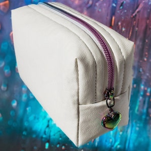May include: A white cosmetic bag with a textured surface and a purple zipper. The zipper pull is a heart-shaped charm with iridescent colors. The bag is rectangular with a slight trapezoid shape.