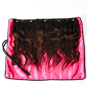 May include: A pink satin hair extension storage bag with a black zipper and a black strap. The bag is open and shows a set of wavy hair extensions.