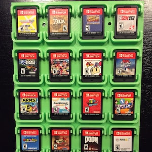 May include: A green plastic wall mount with 16 slots for storing Nintendo Switch game cartridges. The slots are filled with various game cartridges, including "Zelda", "Mario Kart", "Arms", "Doom", and "FIFA 18".