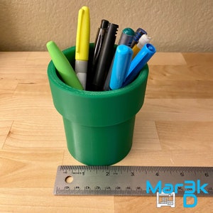 Peut inclure: Un porte-crayons en plastique vert en forme de tuyau. Le porte-crayons est rempli de différents stylos et marqueurs, dont du jaune, du bleu et du noir.
