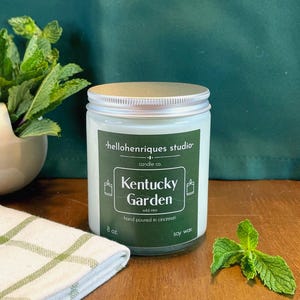 May include: A glass jar candle with a silver lid and a green label that reads "Kentucky Garden" and "wild mint." The candle is 8 oz and hand-poured in Cincinnati. Fresh mint leaves and a white and green striped towel are next to the candle.