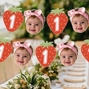 May include: A decorative party banner featuring alternating cutouts of a smiling baby wearing a pink bow headband and a red strawberry shape with a white number one printed on the front, all strung together on a thin brown twine string.