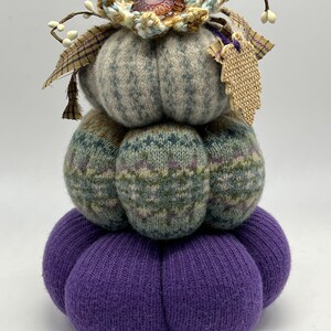 May include: A stack of three knitted pumpkins, the top one is light blue with a crocheted flower on top, the middle one is green and purple with a plaid bow, and the bottom one is purple.