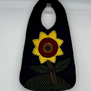 May include: A black felt door hanger featuring a yellow sunflower with a brown center and green leaves. The sunflower has a button in the center. The bottom of the hanger has a green textured design.
