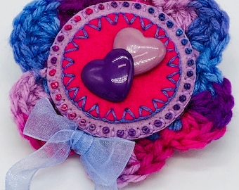 May include: A crocheted flower brooch with a pink, purple, and blue color scheme. The center of the flower features a pink felt circle with two heart-shaped buttons, one purple and one pink.