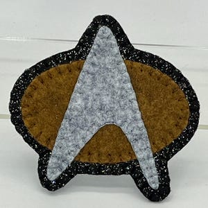 May include: A felt appliqué of the Star Trek insignia, featuring a gold-colored background with a silver-colored star shape. The star shape is outlined in black thread. The background is surrounded by a black border with silver glitter.