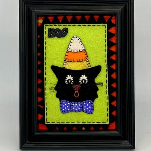 May include: Framed Halloween artwork featuring a black cat wearing a candy corn hat and a blue polka dot bow tie. The cat is set against a lime green background with the word "BOO" above it. The frame is black.