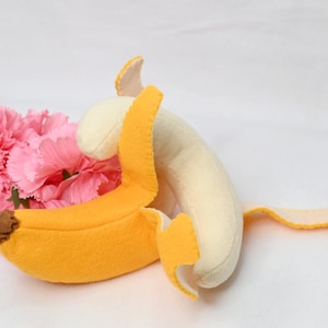May include: A plush banana toy with a yellow peel and a white inner part. The banana is partially peeled and the peel is folded over the banana. The toy is lying on a white surface with pink carnations in the background.