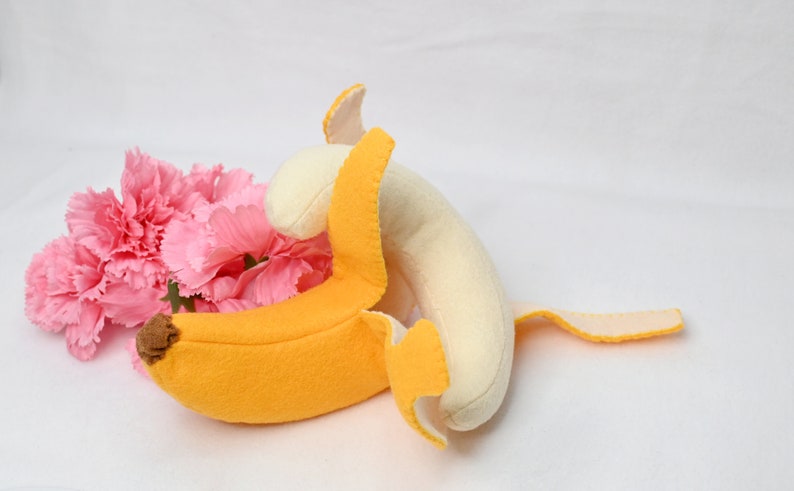 May include: A plush banana toy with a yellow peel and a white inner part. The banana is partially peeled and the peel is folded over the banana. The toy is lying on a white surface with pink carnations in the background.