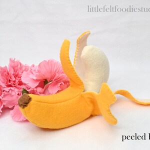 May include: A felt banana toy with a yellow peel and a white inner peel. The banana is peeled and the peel is curled around the banana. The banana is lying on a white surface with pink carnations.