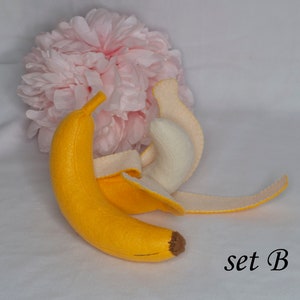 May include: A felt banana toy with a peel that can be removed. The banana is yellow and the peel is cream colored. The toy is set against a white background with a pink flower.