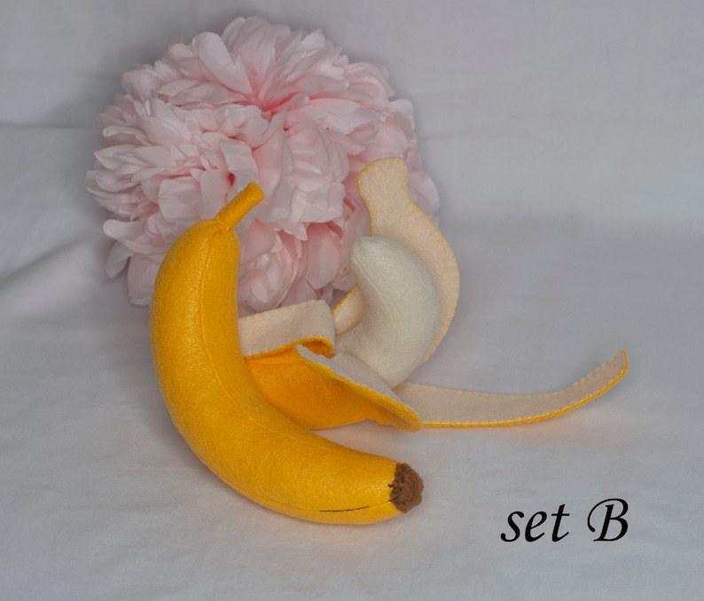 May include: A felt banana toy with a peel that can be removed. The banana is yellow and the peel is cream colored. The toy is set against a white background with a pink flower.