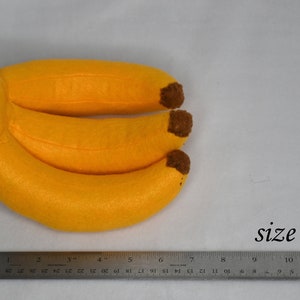 May include: Three yellow felt bananas with brown felt stems, measured against a ruler, showing they are approximately 7 inches long.