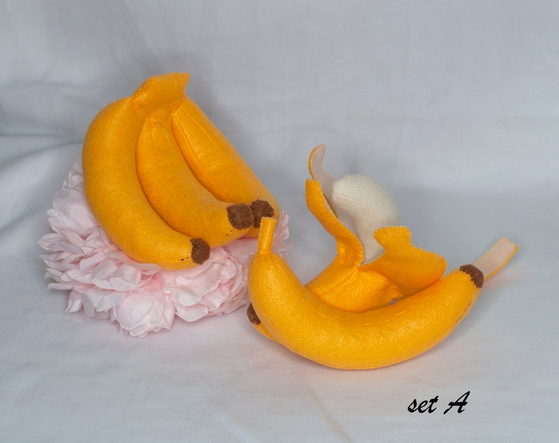 May include: A set of felt banana toys, including a bunch of three bananas and a single banana with a peel. The bananas are yellow with brown tips and the peel is white. The toys are arranged on a pink flower.