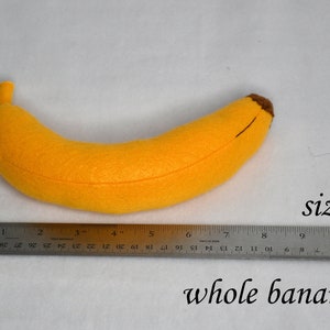 May include: A yellow felt banana plush toy measuring 7 inches long. The banana is lying on a white surface with a ruler next to it.