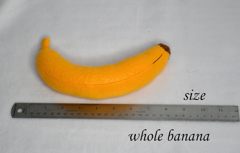 May include: A yellow felt banana plush toy measuring 7 inches long. The banana is lying on a white surface with a ruler next to it.