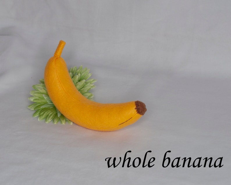 May include: A felt banana toy, yellow with a brown tip, on a white background with green leaves.