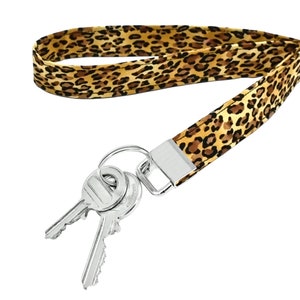 Pode incluir: Um cordão para chaves com estampa de leopardo em tons de castanho e bege. O cordão está preso a um fecho de metal prateado que segura duas chaves prateadas. As chaves têm um formato padrão.