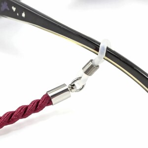 May include: A close-up of a burgundy rope eyeglass strap attached to a pair of glasses with a spring-loaded clasp. The glasses have a black frame with a gold trim and a butterfly design on the temple.