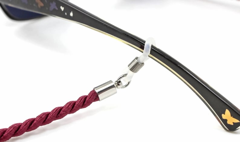 May include: A close-up of a burgundy rope eyeglass strap attached to a pair of glasses with a spring-loaded clasp. The glasses have a black frame with a gold trim and a butterfly design on the temple.