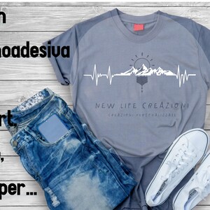 Op de afbeelding: Een grijs T-shirt met een witte bergketen en zon-afbeelding met een hartslaglijn. De tekst "NEW LIFE CREAZIONI" en "CREAZIONI PERSONALIZZATE" staat onder de afbeelding.