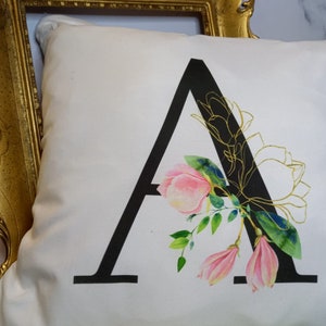 Puede incluir: Una almohada blanca con una letra A negra y un dise&ntilde;o floral rosa y verde. La letra A est&aacute; delineada en oro.
