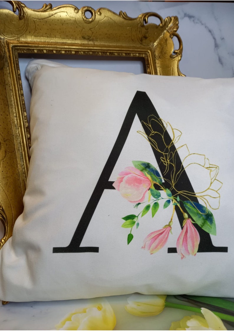 Puede incluir: Una almohada blanca con una letra A negra y un dise&ntilde;o floral rosa y verde. La letra A est&aacute; delineada en oro.