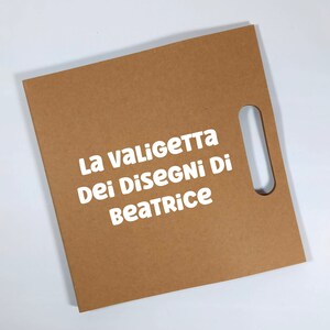 Peut inclure: Un portfolio en carton brun avec une d&eacute;coupe rectangulaire pour la poign&eacute;e. Le texte "La Valigetta Dei Disegni Di Beatrice" est imprim&eacute; en blanc. Le portfolio est sur fond blanc.