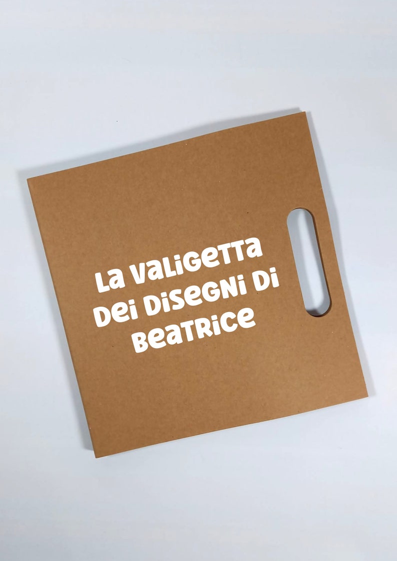 Peut inclure: Un portfolio en carton brun avec une d&eacute;coupe rectangulaire pour la poign&eacute;e. Le texte "La Valigetta Dei Disegni Di Beatrice" est imprim&eacute; en blanc. Le portfolio est sur fond blanc.